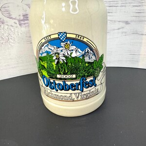 May include: A beige ceramic beer stein with a colorful design. The design includes the words "Oktoberfest Richmond, Virginia" and the year "2002". The stein also features a mountain and flower scene.