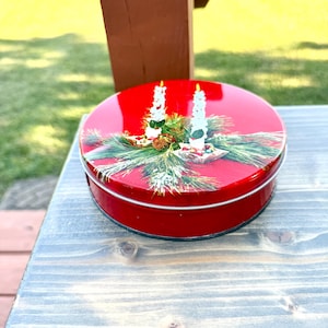 May include: A round red metal tin featuring a festive illustration on the lid. The design depicts two white candles surrounded by green pine needles, small red berries, and a pinecone, creating a holiday-themed decorative container.