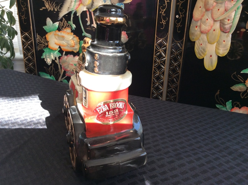May include: A red, black, and white ceramic decanter shaped like a vintage steam engine. The decanter features a gold label with the text "Ezra Brooks 152" and the words "Kentucky Straight Bourbon Whiskey".