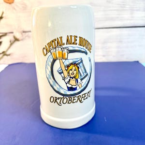 May include: White ceramic beer stein with the words "CAPITAL ALE HOUSE OKTOBERFEST" printed on the front. The stein features a cartoon image of a woman holding beer steins. The stein is cylindrical and has a handle.