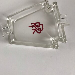 May include: A clear glass ashtray with a red "RK" logo in the center. The ashtray is shaped like a house with a pointed roof and two small holes on the sides.