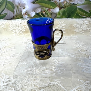 May include: A cobalt blue glass shot glass with a gold metal rim and handle. The glass has a gold metal band around the middle with the word "FREEPORT" inscribed on it.