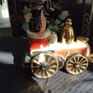 May include: A red, white, and black ceramic train engine decanter with gold accents. The engine has a black chimney and a gold-colored bottle stopper. The decanter is sitting on a dark gray surface.
