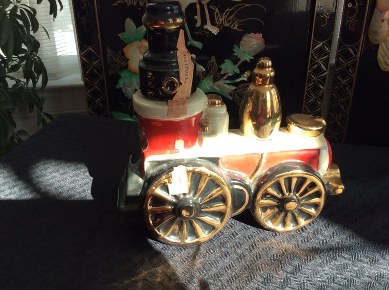 May include: A red, white, and black ceramic train engine decanter with gold accents. The engine has a black chimney and a gold-colored bottle stopper. The decanter is sitting on a dark gray surface.