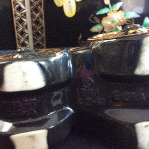 May include: Close-up of two black ceramic teapots with white accents. The teapots have a textured surface and are marked with the text "1971 Ezra Imports".