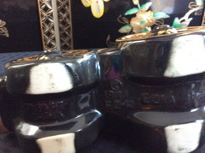 May include: Close-up of two black ceramic teapots with white accents. The teapots have a textured surface and are marked with the text "1971 Ezra Imports".