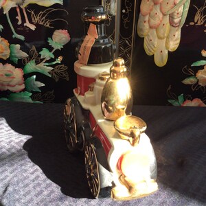 May include: A white ceramic fire engine decanter with gold accents. The decanter features a black and red design and a gold-colored bottle stopper. The decanter is sitting on a black and white patterned surface.