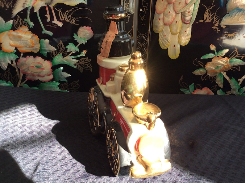 May include: A white ceramic fire engine decanter with gold accents. The decanter features a black and red design and a gold-colored bottle stopper. The decanter is sitting on a black and white patterned surface.