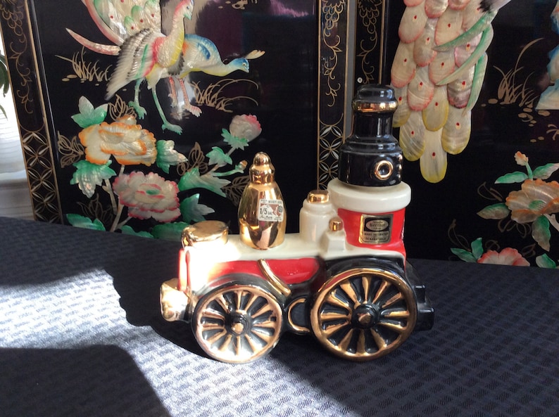 May include: A red, white, and black ceramic train engine decanter with gold accents. The engine has a black chimney and a gold-colored bottle on top. The decanter is sitting on a gray surface.