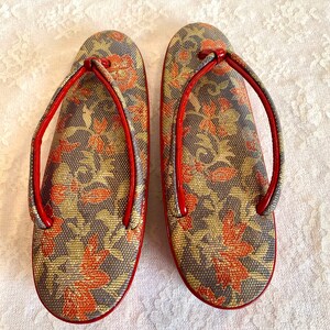 May include: A pair of red and gold Japanese sandals with a floral pattern. The sandals have a raised wooden platform and a strap that goes around the ankle.