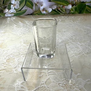 May include: A clear glass square vase with a decorative etched design sits on a clear acrylic square pedestal.
