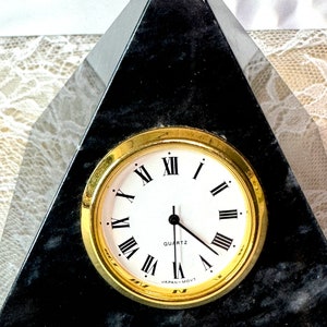 May include: A black marble pyramid clock with a gold-rimmed clock face and Roman numeral markings. The clock face reads "QUARTZ" and "JAPAN MOV'T".