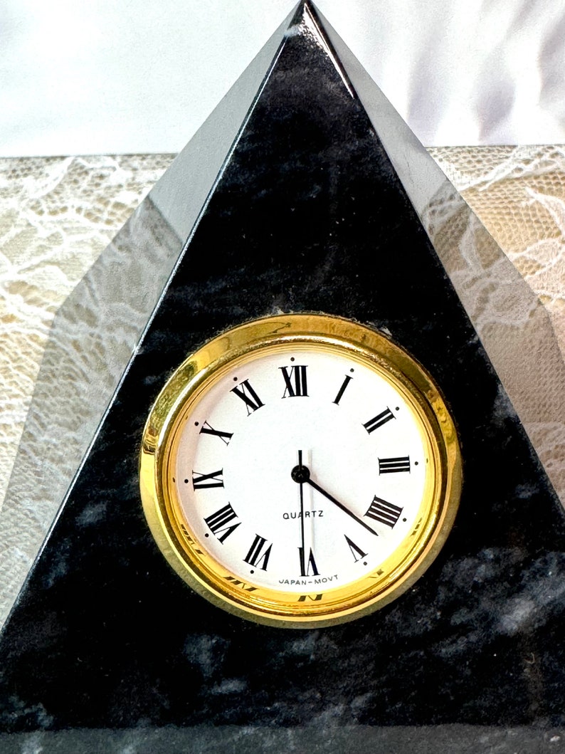 May include: A black marble pyramid clock with a gold-rimmed clock face and Roman numeral markings. The clock face reads "QUARTZ" and "JAPAN MOV'T".