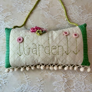 May include: A white pillow with green gingham trim and white pom-poms along the bottom edge. The pillow has the word "Garden" embroidered on it and three pink buttons. A green ribbon loop is attached to the top of the pillow.