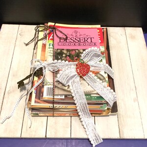 May include: A stack of cookbooks tied with a white ribbon and a red strawberry embellishment. The top cookbook is pink and reads "DESSERT COOKBOOK". A decorative key and a black and gold chain are also tied to the stack.