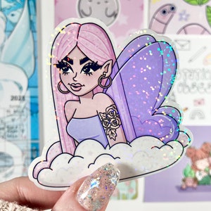 May include: A holographic sticker featuring a cartoon fairy with pink hair, purple wings, and a floral tattoo on her arm. She is sitting on a cloud with a sparkly background.