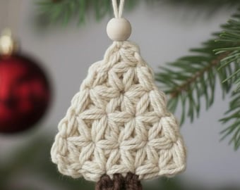 Puede incluir: Adorno de árbol de Navidad de ganchillo color crema con tronco marrón. El árbol está suspendido por una cuerda blanca y una cuenta de madera. Una bola de Navidad roja está borrosa en el fondo, sobre un pino verde.