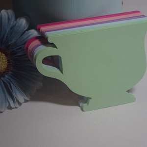 May include: A stack of green, pink, purple, and blue paper teacup shapes. The teacups are cut out and ready to be used for crafts or decorations.