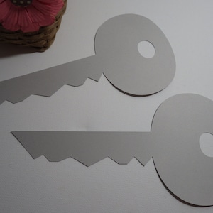 May include: Two gray paper keys with a jagged edge on the bottom. The keys are on a white surface.