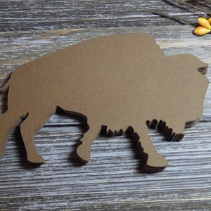 May include: A stack of brown paper cutouts in the shape of a bison. The bison is in profile, walking to the left. The background is a weathered, gray wooden surface. Decorative sprigs with orange and yellow berries are in the upper right corner.