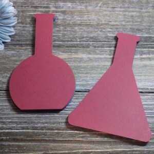 May include: Two red paper cutouts of beakers on a wood background. The beakers are different shapes, one is round and the other is a triangle.