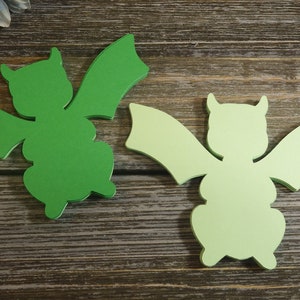 May include: Two green paper cutouts of a dragon with wings and horns. The dragon on the left is a darker green, while the dragon on the right is a lighter green.