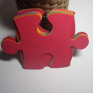 K&ouml;nnte beinhalten: Ein Stapel roter Puzzle-Teile mit regenbogenfarbenen Kanten. Die Teile sind aus dickem Papier oder Karton.