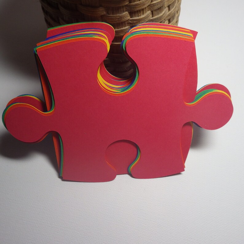 K&ouml;nnte beinhalten: Ein Stapel roter Puzzle-Teile mit regenbogenfarbenen Kanten. Die Teile sind aus dickem Papier oder Karton.