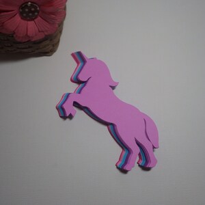 May include: A layered paper unicorn cutout in shades of pink, purple, and blue. The unicorn is standing on its hind legs with its head tilted back.