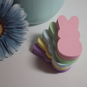 May include: A stack of pastel colored paper bunny shapes. The bunny shapes are layered in a rainbow pattern, starting with purple, then yellow, light blue, green, and pink on top.