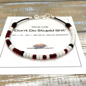 May include: Red, white, and black beaded bracelet with a black leather cord and silver clasp. The bracelet spells out "Don't Do Stupid Shit" in Morse code.