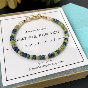 May include: A beaded bracelet featuring small, round, mottled blue and green stones interspersed with white beads, finished with a gold-toned metal clasp. The bracelet rests on a white card printed with Morse Code, the text GRATEFUL FOR YOU, and the URL SunsetSouthpaw.etsy.com.