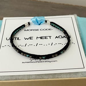 May include: A black beaded bracelet with teal and gray accents, featuring a light blue ribbon and silver-toned metal ends. The card reads "MORSE CODE - UNTIL WE MEET AGAIN" with Morse code symbols.