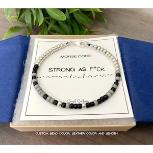 May include: A silver beaded bracelet with black and gray beads spelling "STRONG AS F*CK" in Morse code. The bracelet is displayed in a gift box with a card that reads "CUSTOM BEAD COLOR, LEATHER COLOR AND LENGTH".