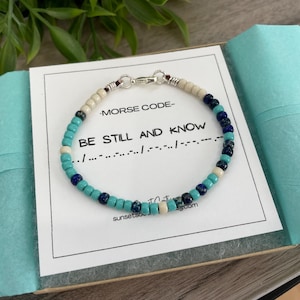 May include: A turquoise, blue and white beaded bracelet with a silver clasp. The bracelet spells out "BE STILL AND KNOW" in Morse code.