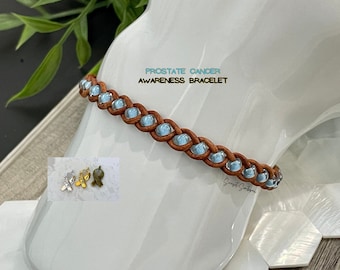 May include: A brown leather bracelet with light blue beads and a braided design. The bracelet has a charm with the word "HOPE" on it. The text "PROSTATE CANCER AWARENESS BRACELET" is written on a white background.