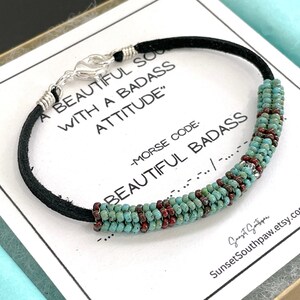 May include: A black leather bracelet with turquoise and red beads. The bracelet has a silver clasp and the text "A BEAUTIFUL SO WITH A BADASS ATTITUDE" and "MORSE CODE: BEAUTIFUL BADASS" is printed on the card.