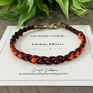 May include: Braided brown leather bracelet with orange and red beads. The bracelet is on a white card with the text "Morse Code - Hokie Mom". The card also includes the Etsy shop's URL.