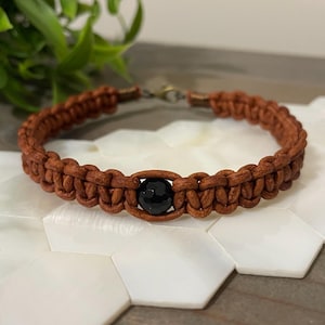 May include: A brown macrame bracelet with a black bead in the center. The bracelet is made of woven cord and has a bronze-colored clasp. The bracelet is displayed on a white, hexagonal surface.