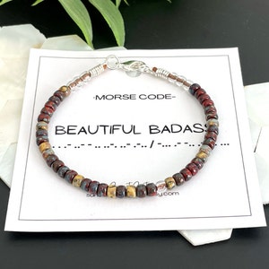 May include: A beaded bracelet with red, brown, and gold beads. The bracelet has a silver clasp and is on a white card with the text "Morse Code - Beautiful Badass" and a code in Morse code.