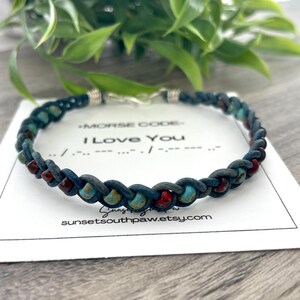 May include: A blue braided leather bracelet with red and turquoise beads. The bracelet has a silver clasp and is on a white card with the text "Morse Code - I Love You" and a website address.
