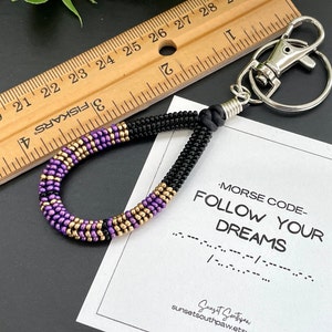 Peut inclure: Porte-clés en perles violettes, dorées et noires avec un fermoir à homard argenté et un petit anneau argenté. Le porte-clés est attaché à une carte avec le texte "MORSE CODE- FOLLOW YOUR DREAMS" et la traduction en code morse de la phrase.
