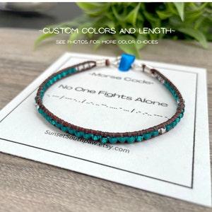 May include: Teal blue beaded bracelet with brown leather cord and silver clasp. The bracelet has the message "No One Fights Alone" in Morse code.