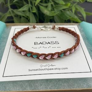 May include: A brown leather bracelet with pink and turquoise beads. The bracelet is braided and has a silver clasp. The card in the background says "Morse Code" and "BADASS" in all caps. The card also includes the text "SunsetSouthpaw.etsy.com".