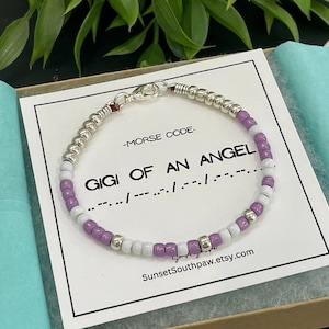 May include: A beaded bracelet featuring alternating purple, white, and metallic silver beads, secured with a silver lobster clasp. The bracelet rests on a white card printed with the text -MORSE CODE- GIGI OF AN ANGEL and Morse code dots and dashes.