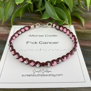 May include: A brown leather bracelet with pink and clear beads. The bracelet is on a white card with the text "Morse Code" and "F*ck Cancer" in black. The card also includes the Morse code for the phrase.