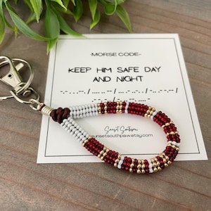 May include: A beaded wristlet keychain with a silver clasp. The wristlet features a pattern of red, white, and gold beads. A card with the text "KEEP HIM SAFE DAY AND NIGHT" and Morse code is in the background.