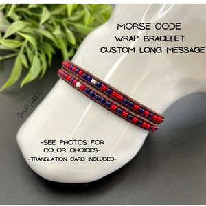 May include: A brown leather wrap bracelet with red, white, and blue beads. The bracelet is on a white mannequin arm. The text "MORSE CODE WRAP BRACELET CUSTOM LONG MESSAGE" is above the bracelet. The text "SEE PHOTOS FOR COLOR CHOICES" and "TRANSLATION CARD INCLUDED" are below the bracelet.