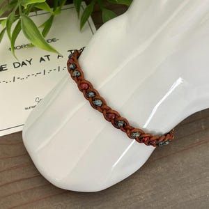 May include: A braided leather bracelet with red and blue-green beads. The bracelet is displayed on a white ceramic form. A card with text that reads "ONE DAY AT A TIME" is in the background.