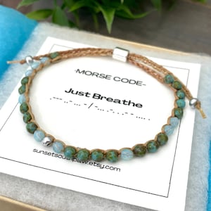 May include: A beaded bracelet with the Morse code message "Just Breathe." The bracelet features green and blue beads strung on a tan cord, with a silver-colored clasp. The bracelet is presented on a white card inside a blue box.